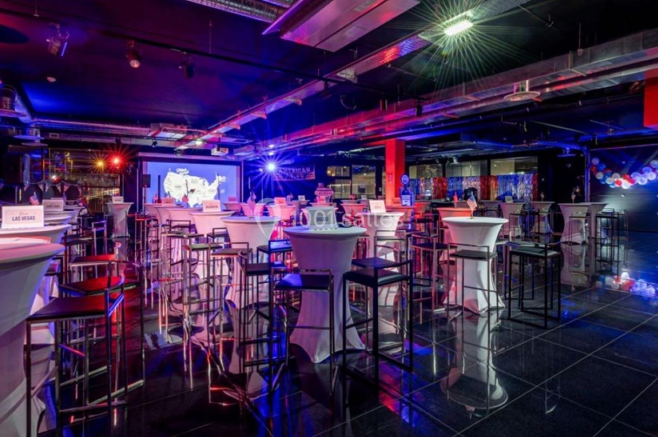 Salle événementielle moderne avec tables hautes, chaises, éclairage coloré et écran de projection en arrière-plan.