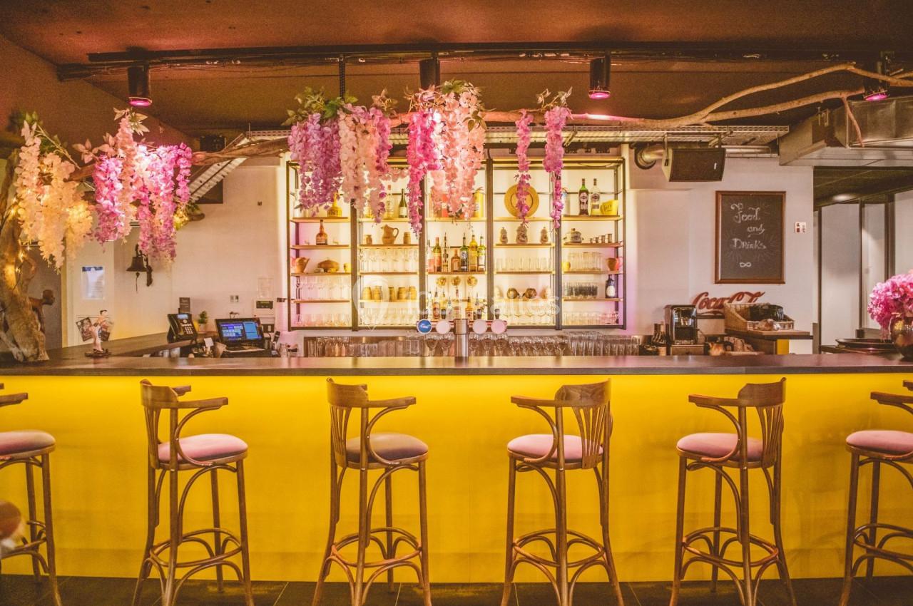 Bar lumineux avec comptoir jaune, tabourets hauts en bois et décorations florales suspendues devant des étagères de…