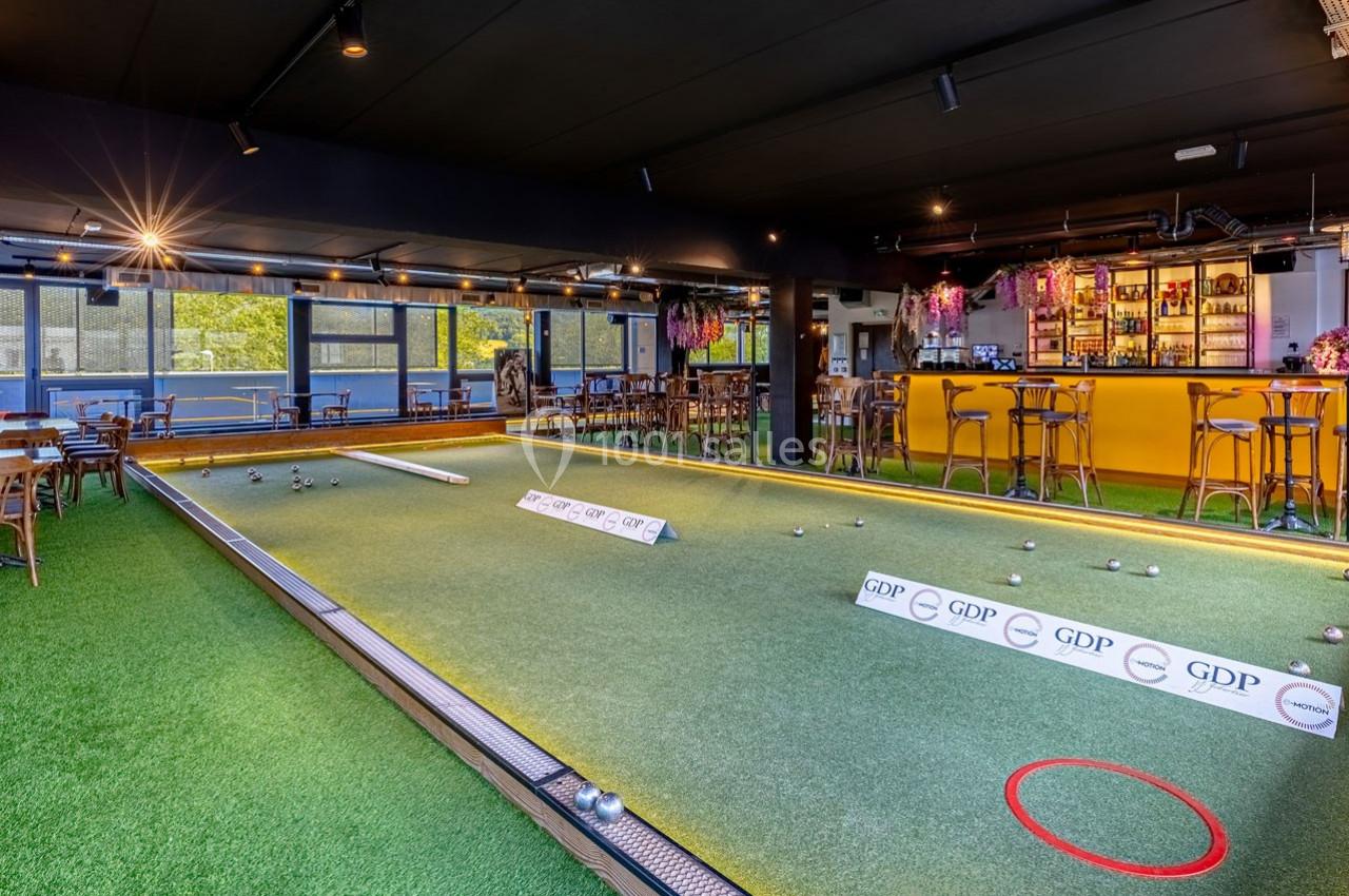 Terrain de pétanque intérieur avec sol en gazon synthétique, entouré de tables et d'un bar éclairé en arrière-plan.