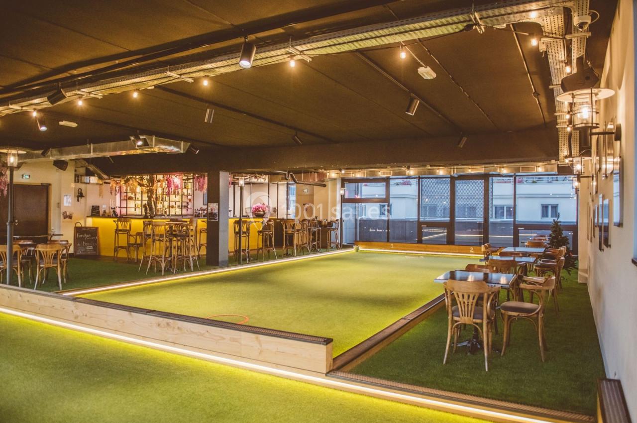 Salle intérieure avec terrain de jeu en gazon synthétique, tables en bois, bar éclairé et grandes fenêtres.