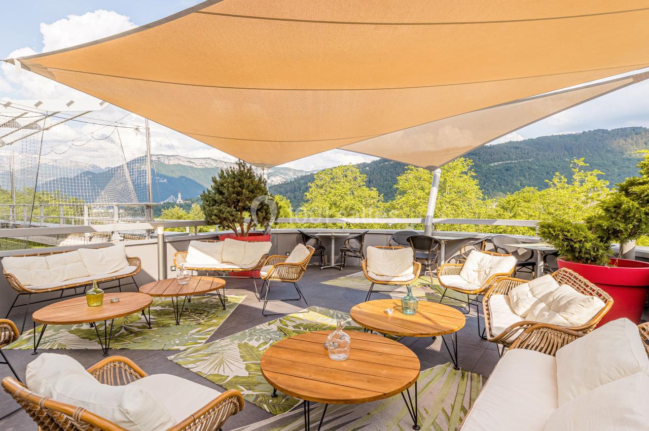 Terrasse aménagée avec des fauteuils en rotin, des tables en bois et des voiles d'ombrage, entourée de montagnes verdoyantes.