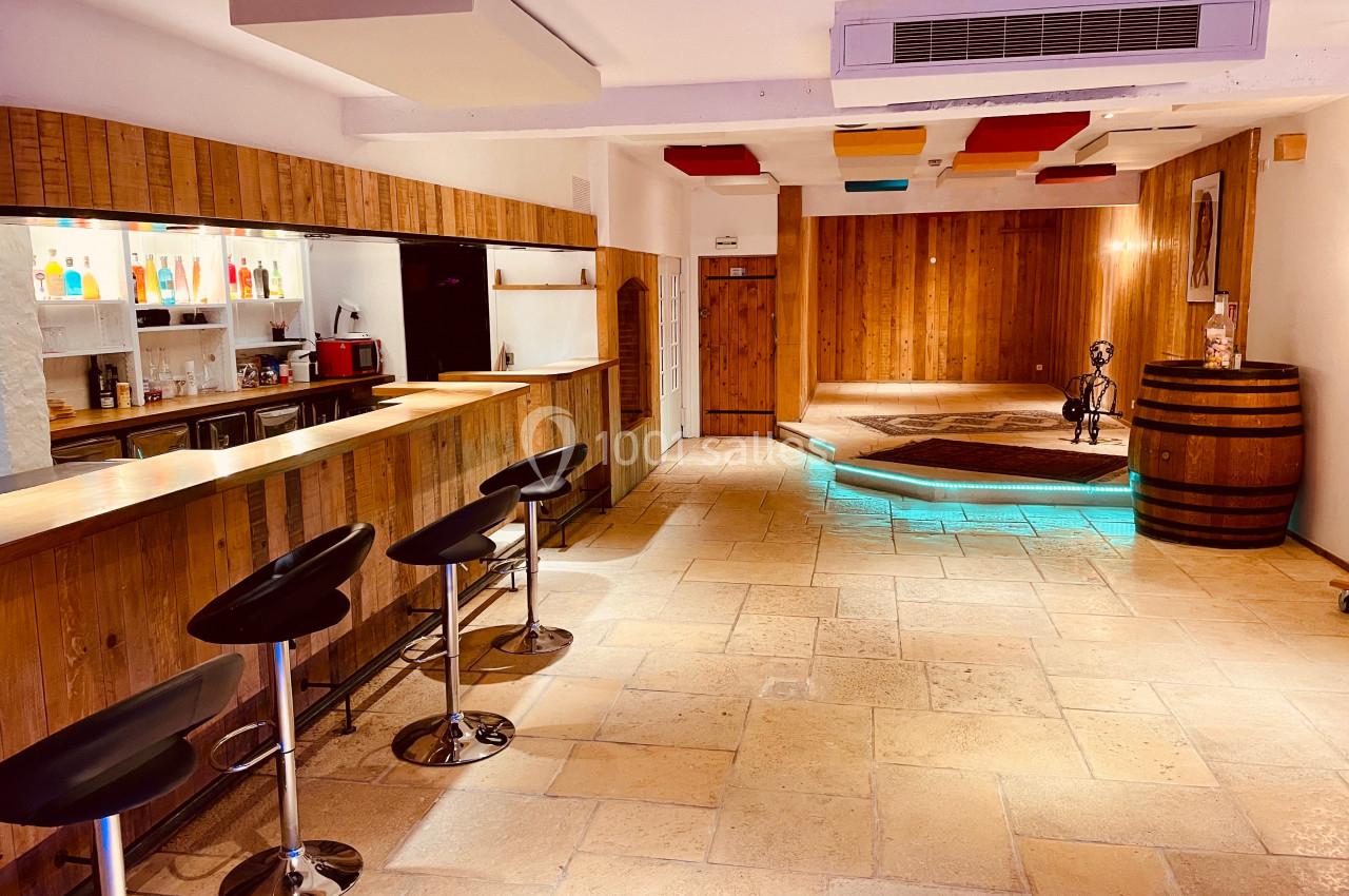 Bar en bois avec tabourets noirs, étagères éclairées de bouteilles colorées et espace lounge avec sol en pierre.