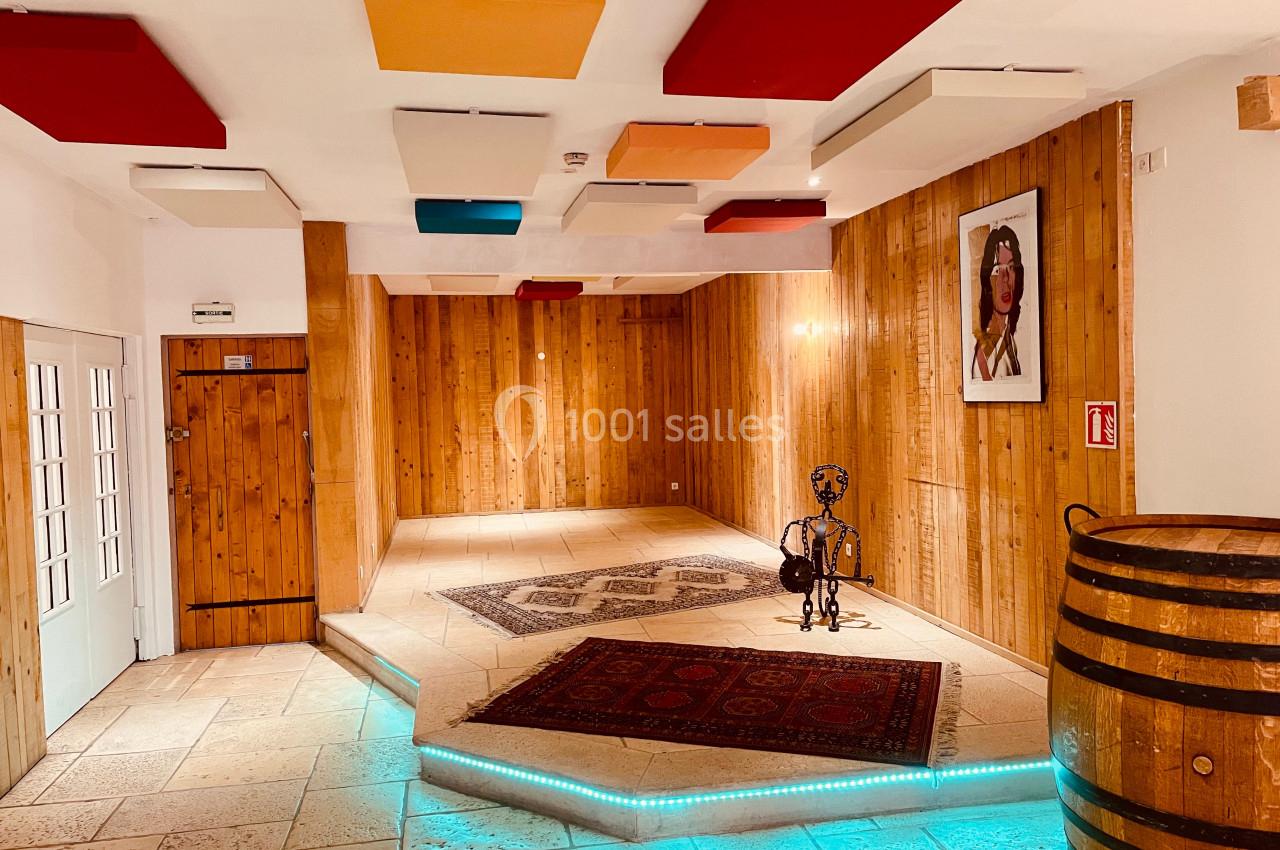 Salle en bois avec scène surélevée, tapis, panneaux acoustiques colorés au plafond et baril décoratif au premier plan.