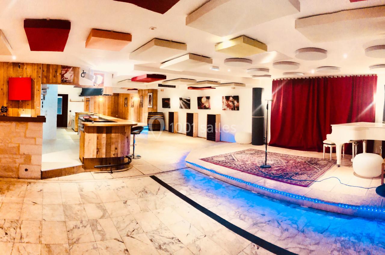 Salle de spectacle avec scène éclairée, micro sur pied, piano blanc et bar en arrière-plan.