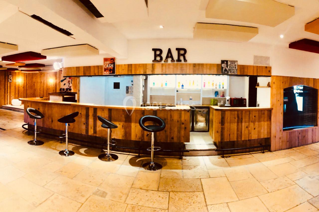 Salle lumineuse avec un bar en bois, tabourets noirs, éclairage coloré et plafond décoré de panneaux acoustiques.