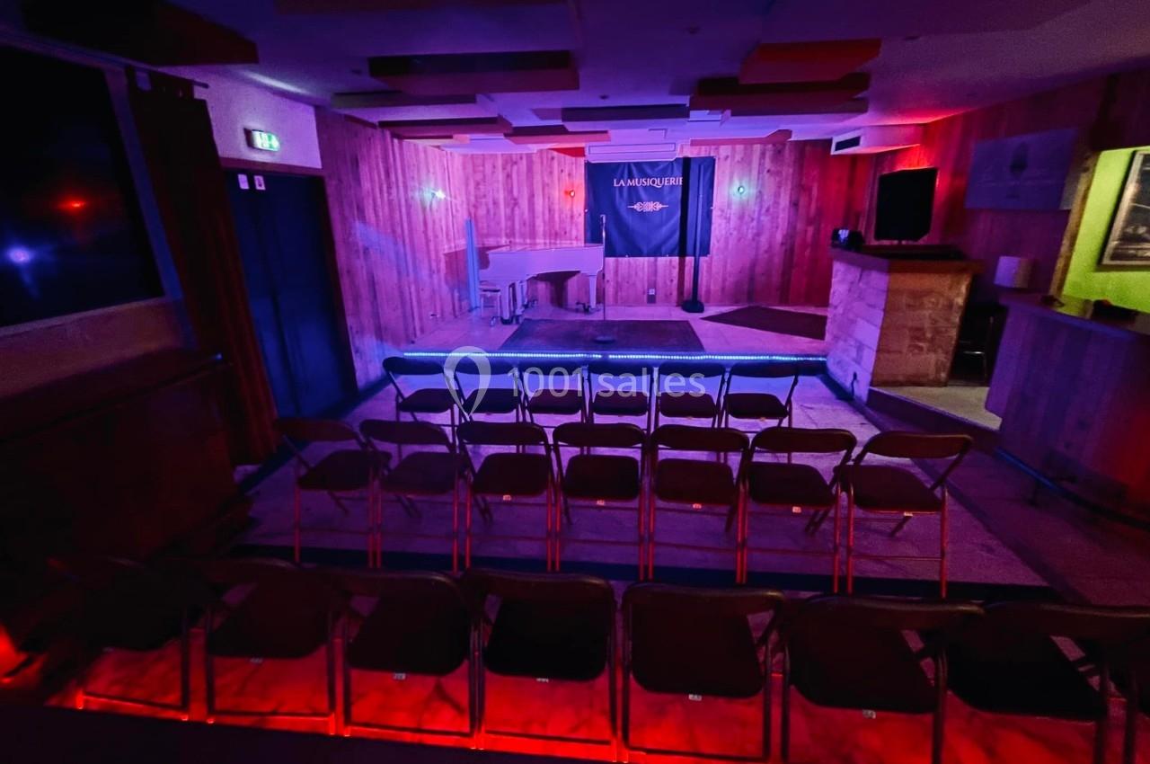 Salle de spectacle avec scène éclairée, piano blanc et rangées de chaises vides dans une ambiance tamisée.