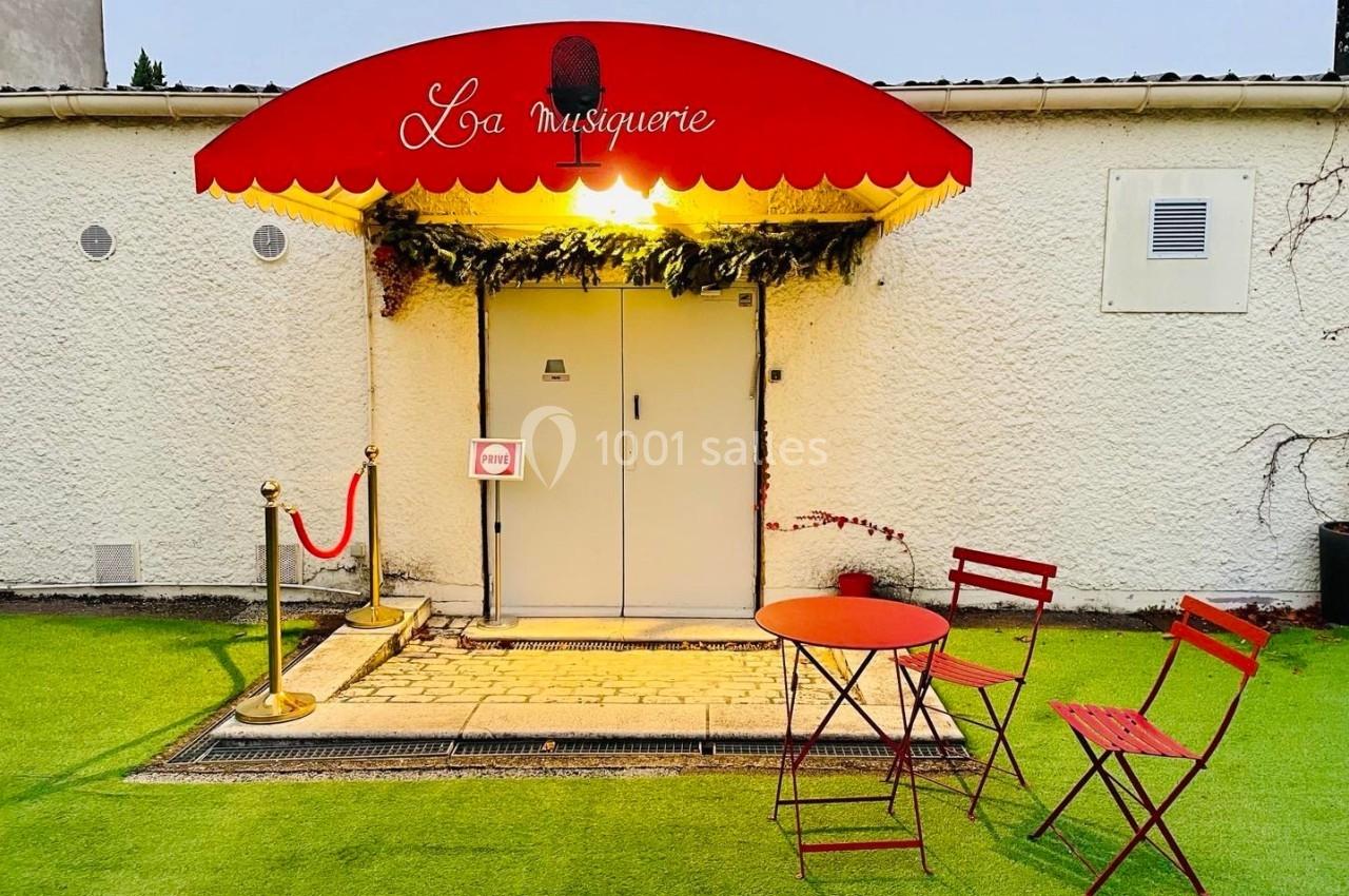Entrée d'un bâtiment avec une enseigne rouge ’La musiquerie’, une table et des chaises rouges sur une pelouse artificielle.