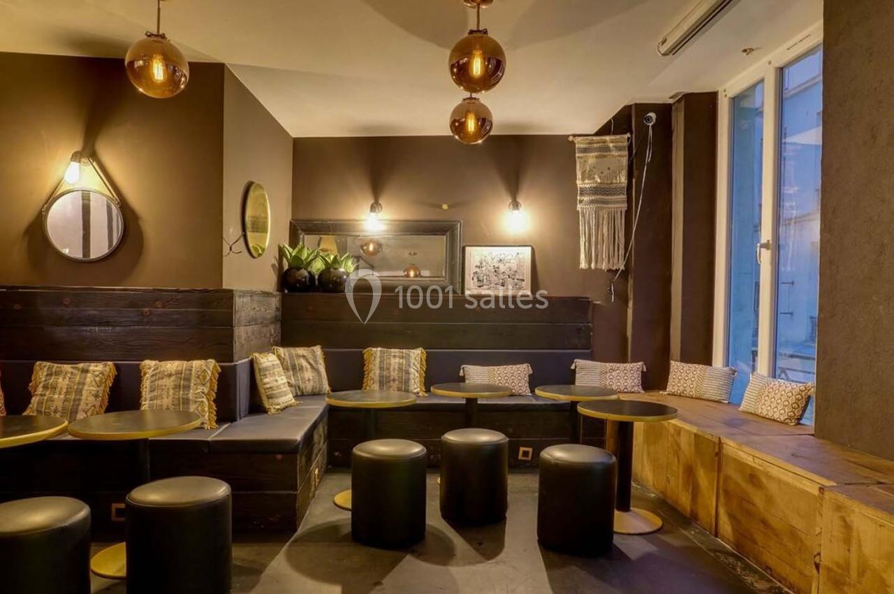 Salon cosy avec banquettes, coussins colorés, tables basses noires et éclairage chaleureux dans une ambiance tamisée.