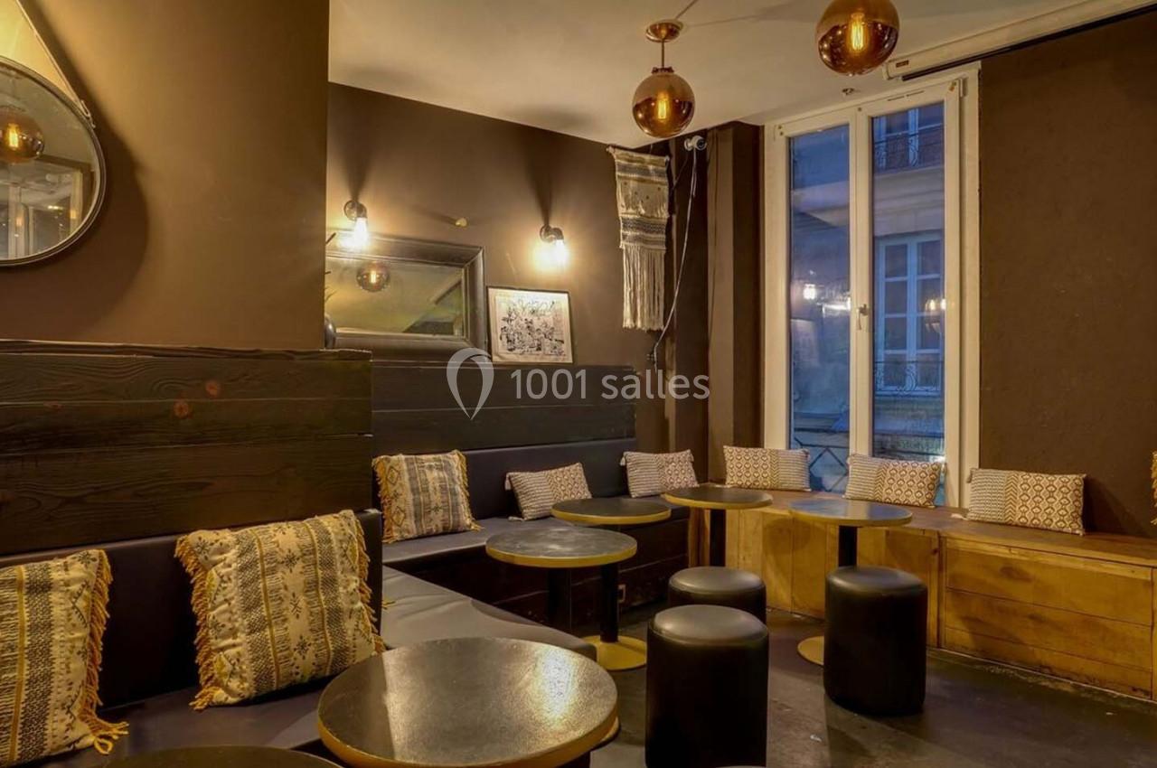 Salon cosy avec tables rondes, tabourets noirs, coussins décoratifs et éclairage tamisé près de grandes fenêtres.