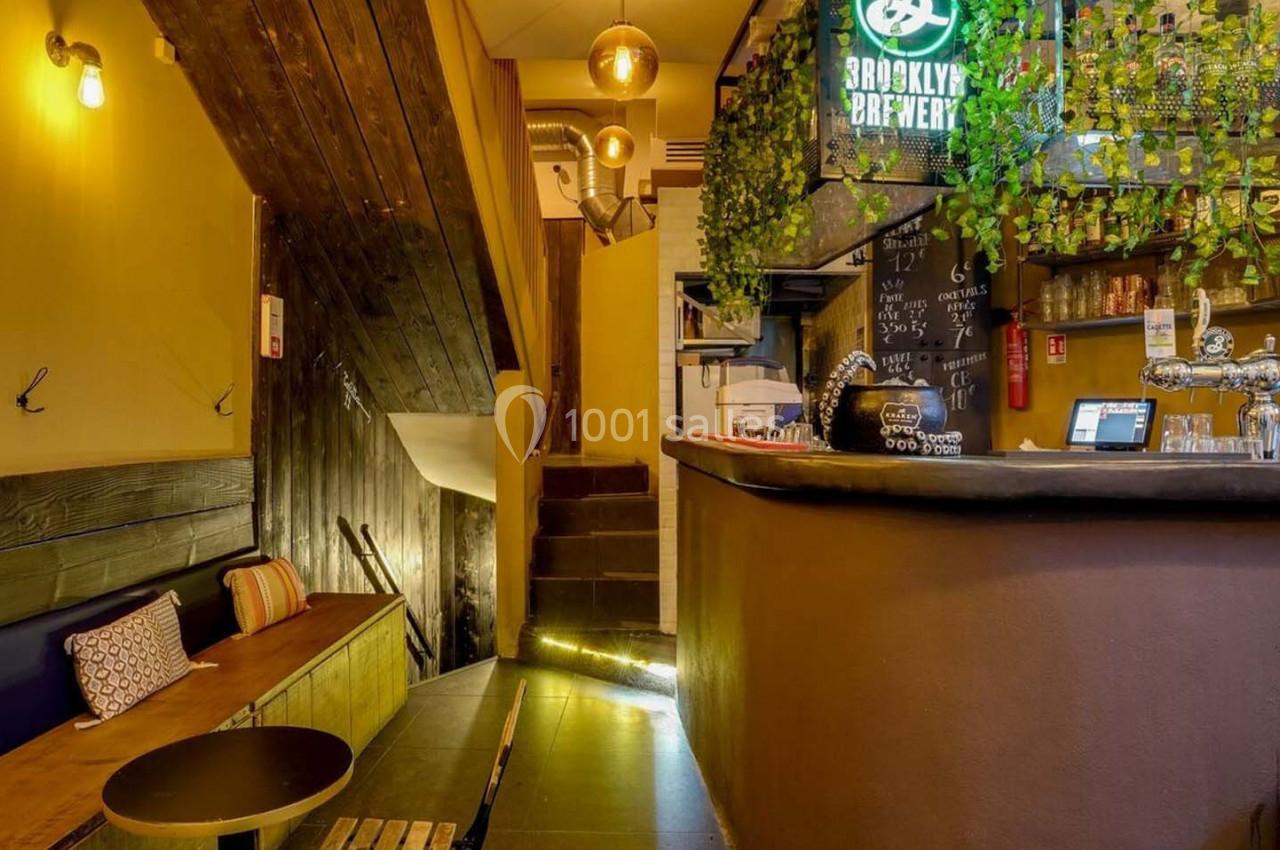 Intérieur d'un bar chaleureux avec un comptoir, des tabourets, des plantes suspendues et un éclairage tamisé.