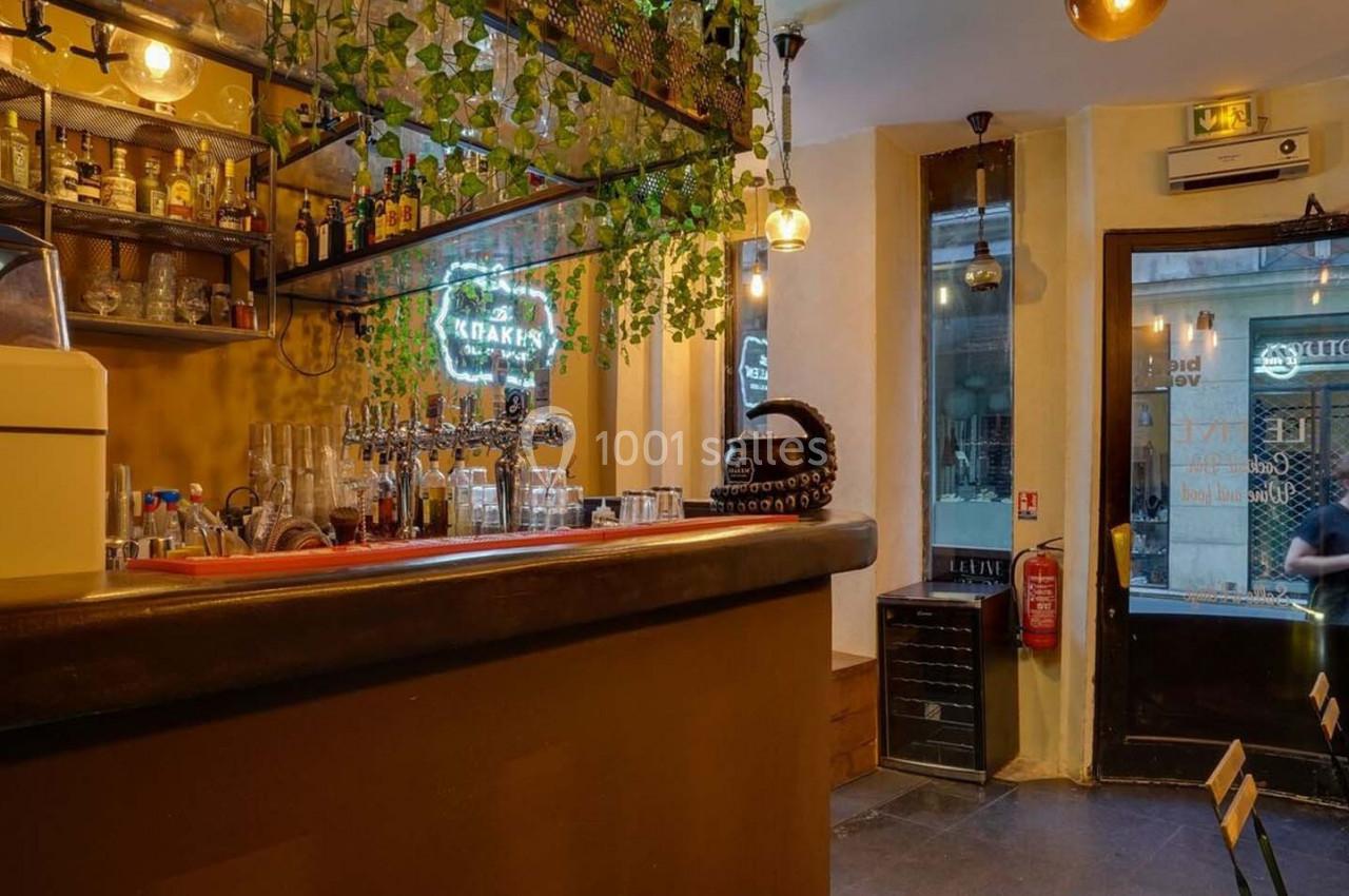Intérieur d'un bar avec comptoir en bois, étagères de bouteilles, plantes suspendues et lumière tamisée.