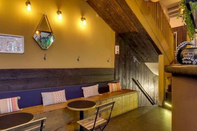 Bar avec comptoir en bois, étagères de bouteilles, plantes suspendues et enseigne lumineuse murale.