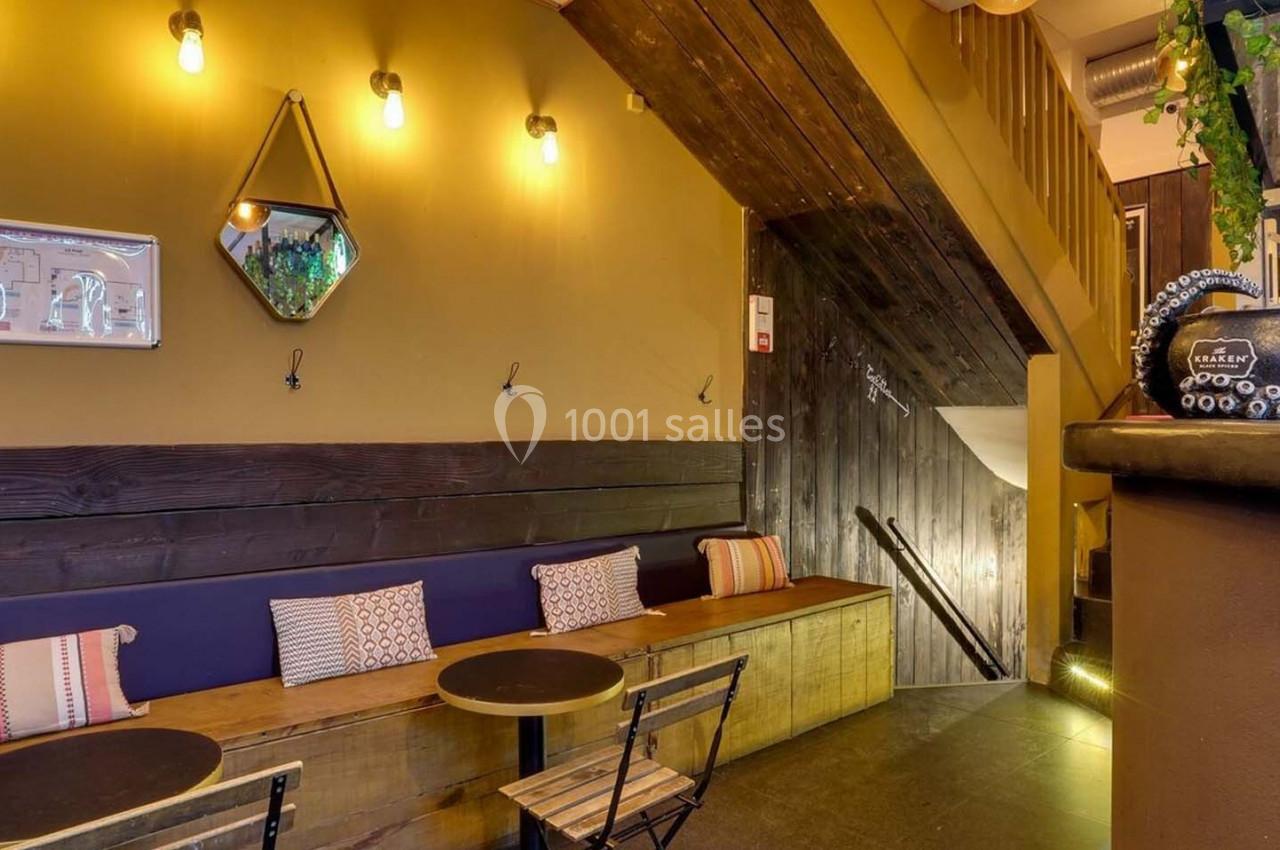 Espace intérieur chaleureux avec banquettes en bois, tables rondes, éclairage doux et escalier en arrière-plan.