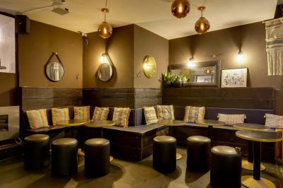 Bar avec comptoir en bois, étagères de bouteilles, plantes suspendues et enseigne lumineuse murale.