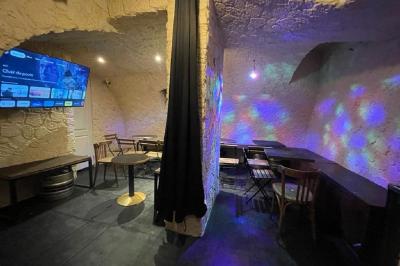Bar avec comptoir en bois, étagères de bouteilles, plantes suspendues et enseigne lumineuse murale.