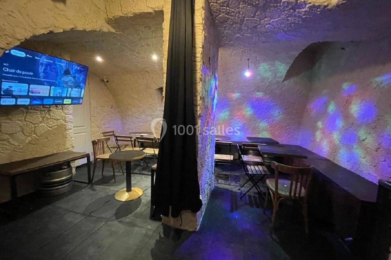 Salle voûtée avec murs en pierre, tables et chaises en bois, éclairage tamisé avec projections lumineuses colorées.