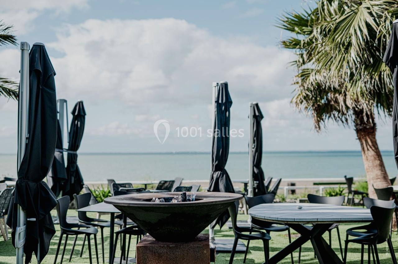 Terrasse avec tables, chaises, parasols fermés, brasero central, vue sur la mer et palmiers sous un ciel partiellement…
