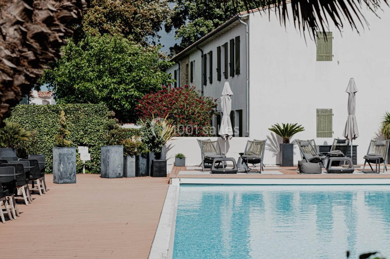 Piscine extérieure entourée de chaises longues, parasols et végétation, avec une maison blanche en arrière-plan.