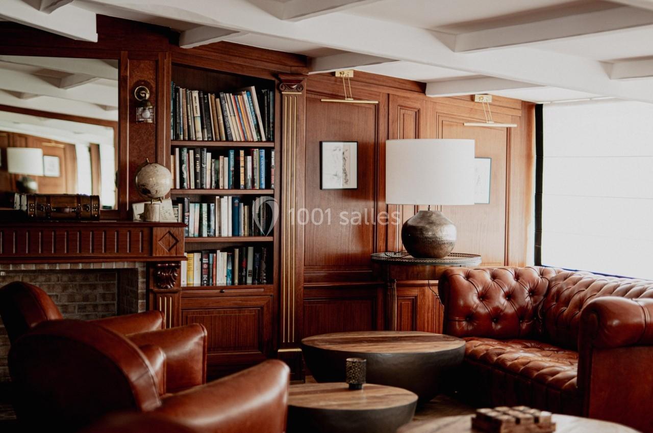 Salon chaleureux avec fauteuils en cuir marron, tables basses en bois, bibliothèque et éclairage naturel.