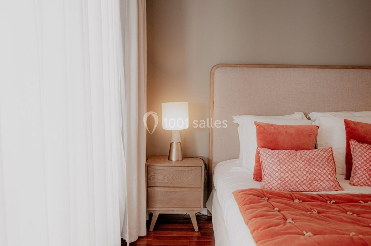 Chambre lumineuse avec lit double, couvre-lit orange, coussins assortis, table de chevet en bois et lampe allumée.