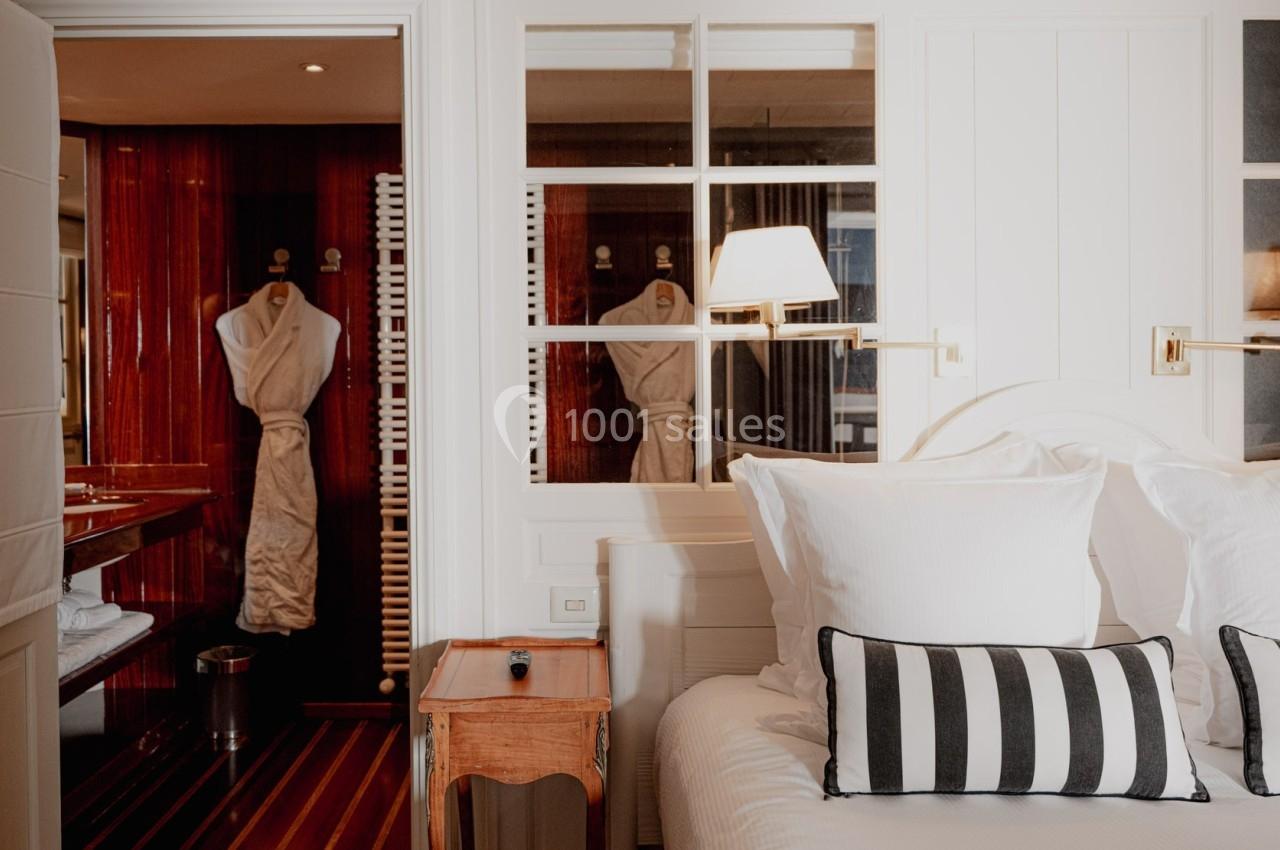 Chambre lumineuse avec lit double, oreillers et plaid rayés, vue sur une salle de bain en bois avec peignoir suspendu.