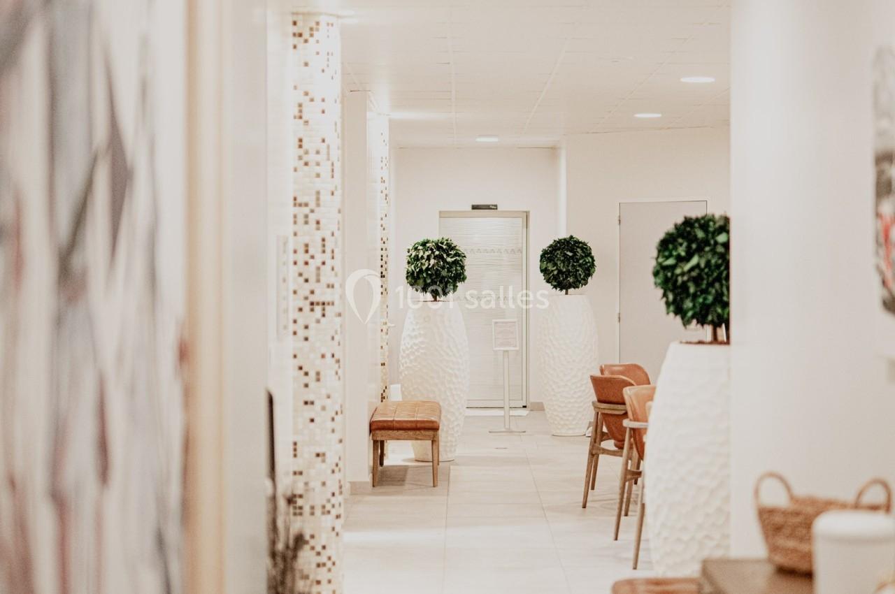 Couloir lumineux avec murs clairs, plantes décoratives en pots blancs, bancs en bois et carrelage beige.