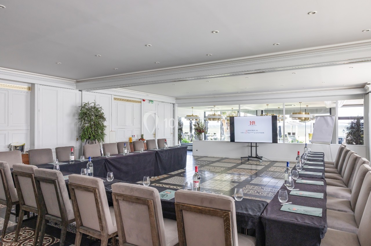 Salle de réunion lumineuse avec tables en U, chaises, bouteilles d'eau, écran de présentation et grandes fenêtres.