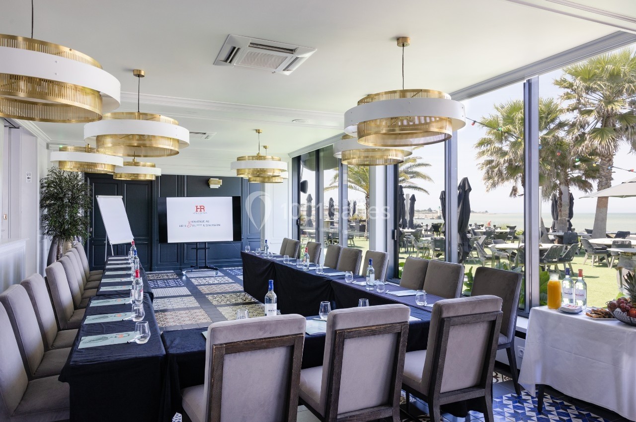 Salle de réunion lumineuse avec une grande table, chaises, écran de présentation et vue sur une terrasse et des palmiers.