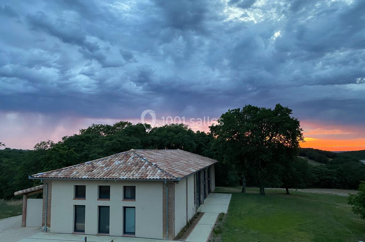 Bâtiment moderne avec toit en tuiles, entouré de verdure, sous un ciel nuageux au coucher du soleil.