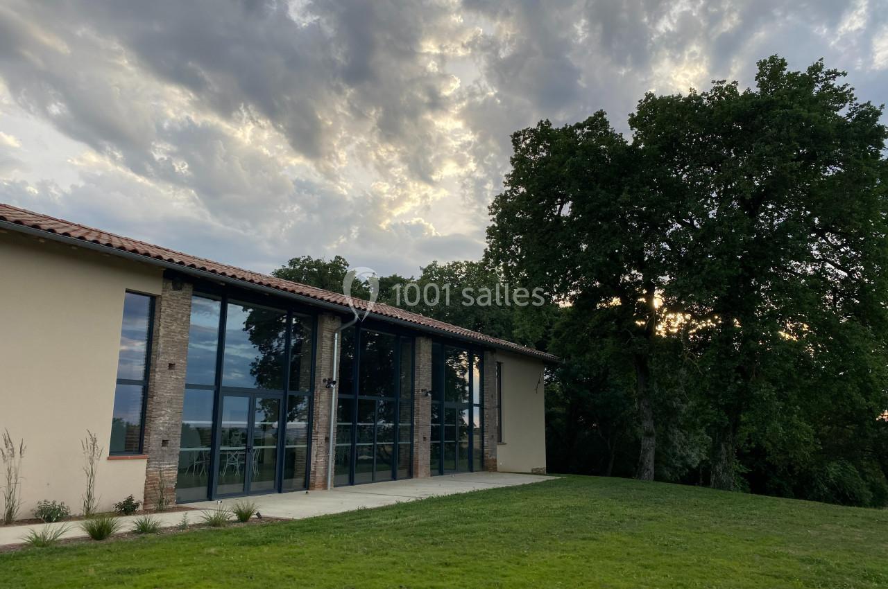Bâtiment moderne avec de grandes baies vitrées, entouré de pelouse et d'arbres sous un ciel nuageux.