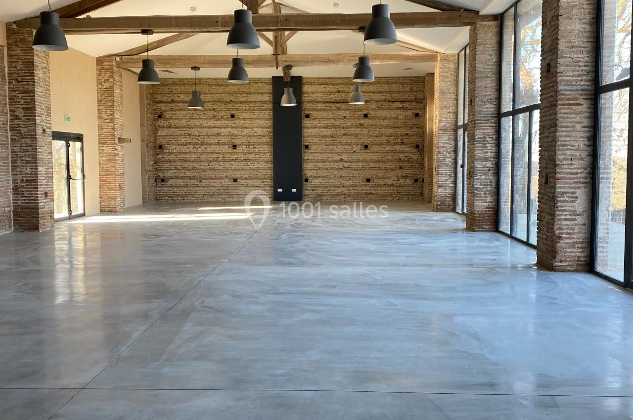 Salle spacieuse avec murs en briques, plafond à poutres apparentes et grandes baies vitrées laissant entrer la lumière…