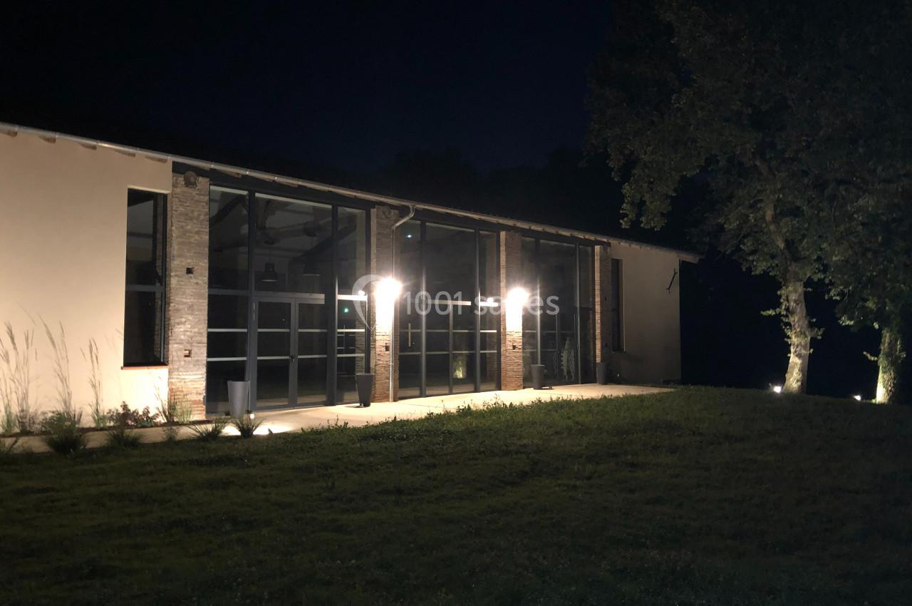 Façade d'un bâtiment moderne éclairé la nuit, avec des arbres et une pelouse au premier plan.