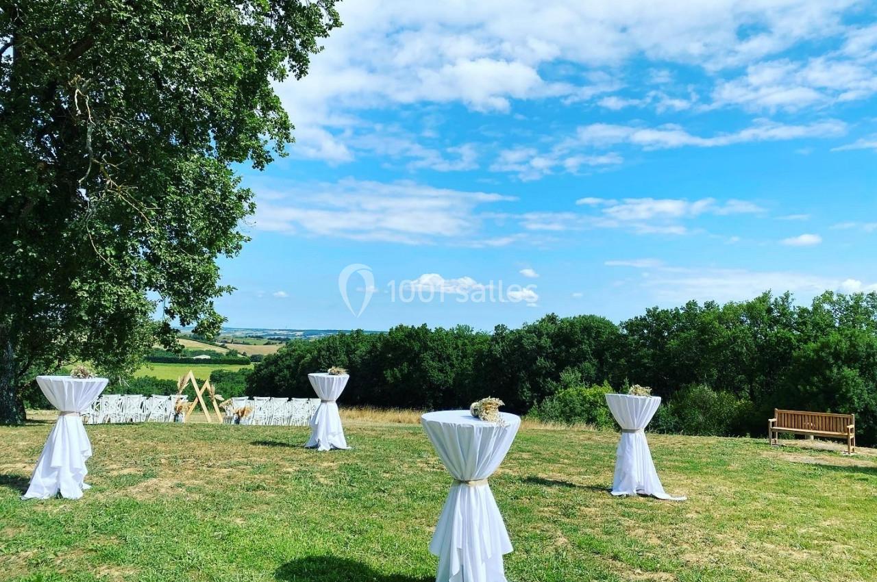 Tables hautes drapées de blanc disposées en extérieur sur une pelouse, avec vue sur un paysage vallonné.