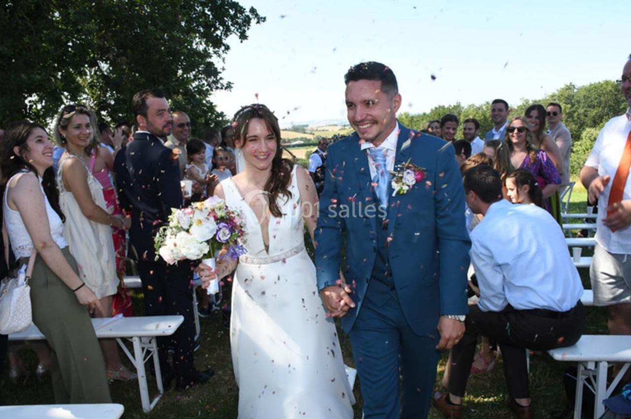 Un couple souriant marche main dans la main sous une pluie de confettis, entouré d'invités lors d'une cérémonie en plein air.