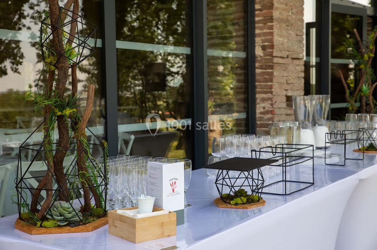 Table décorée avec des éléments végétaux, des verres alignés et des carafes, près d'une fenêtre et d'un mur en briques.