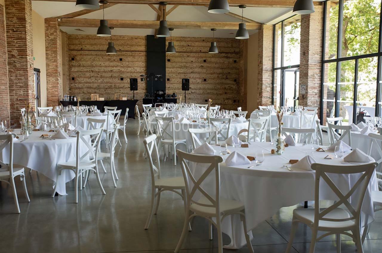 Salle de réception lumineuse avec tables rondes dressées de nappes blanches, chaises en bois et grandes baies vitrées.