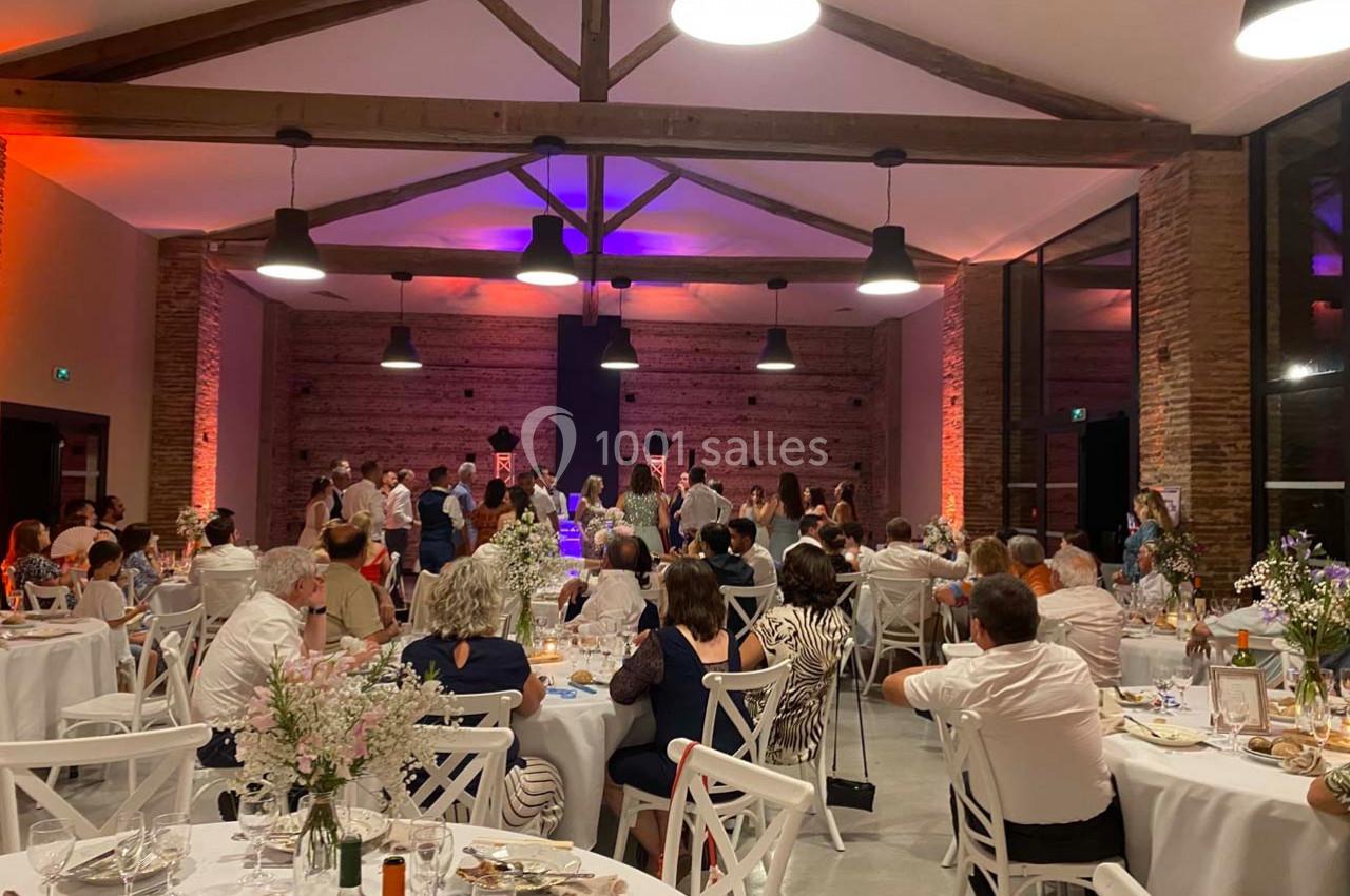 Salle de réception avec des invités assis et debout, éclairée par des lumières colorées et décorée de tables dressées.