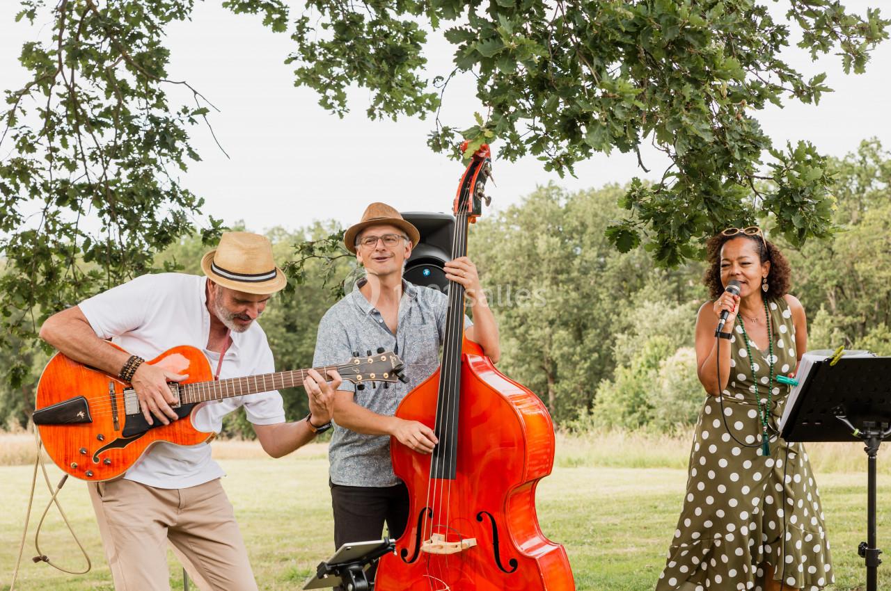 Trois musiciens jouent en plein air : un guitariste, un contrebassiste et une chanteuse sous un arbre.