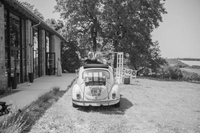 Un couple dans une voiture ancienne quitte un bâtiment entouré de verdure par une journée ensoleillée.