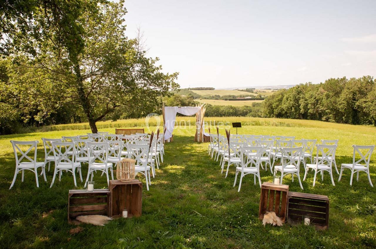 Chaises blanches disposées en extérieur devant une arche décorée, dans un champ verdoyant sous un ciel dégagé.