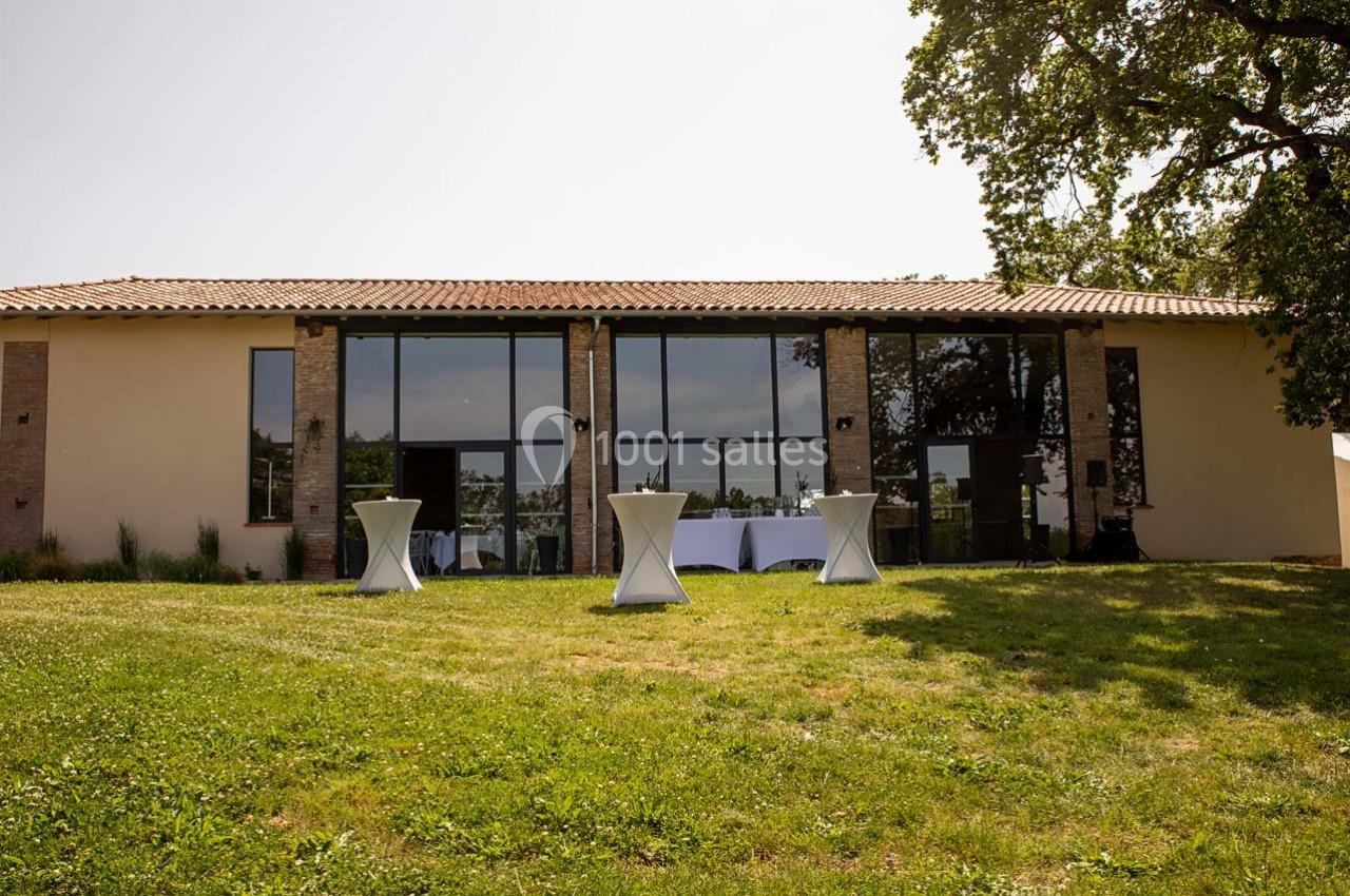 Façade d'un bâtiment moderne avec de grandes baies vitrées, entouré d'herbe et d'arbres, tables hautes à l'extérieur.