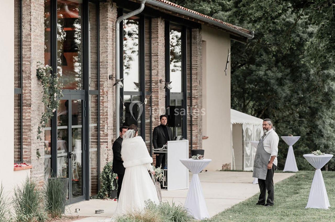 Une mariée en robe blanche se tient devant un bâtiment avec des baies vitrées, entourée de quelques personnes.