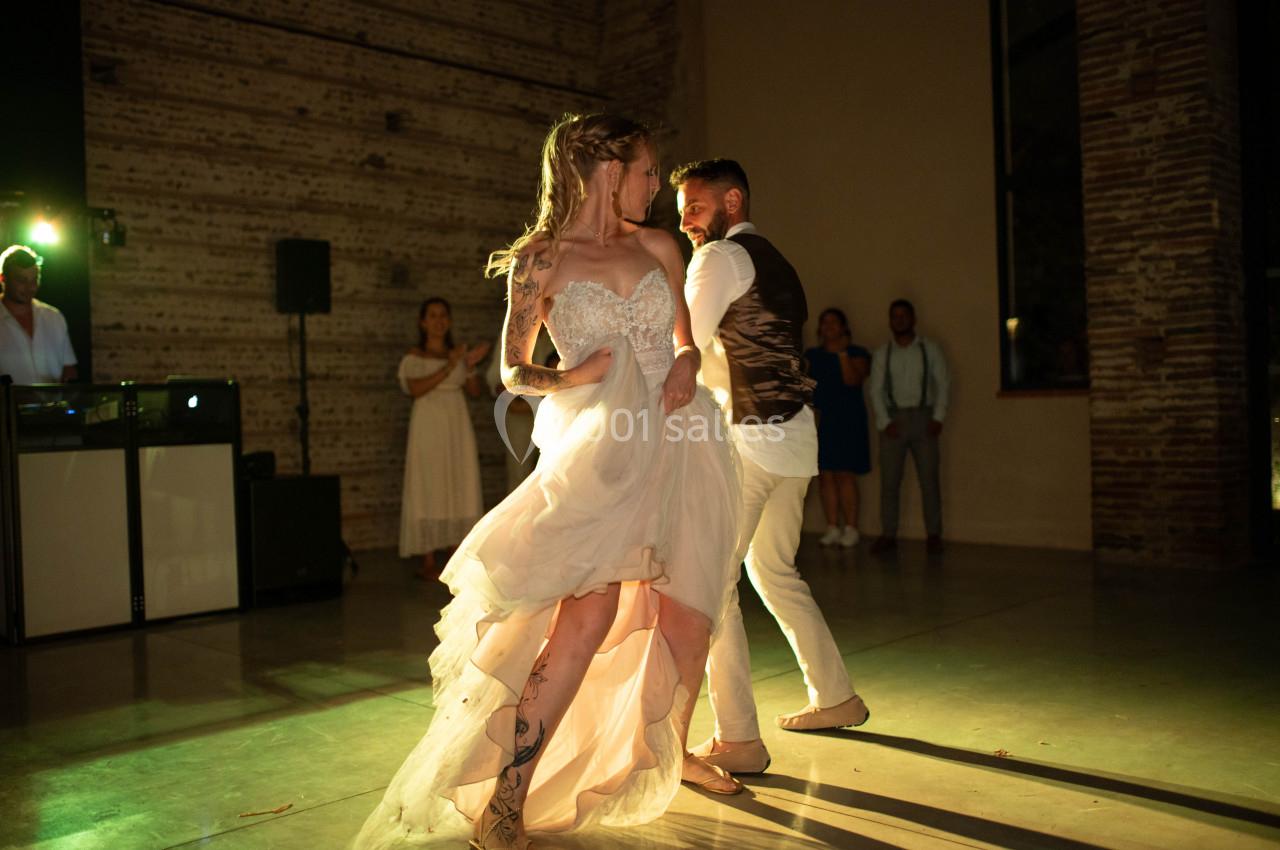 Un couple danse dans une salle éclairée, entouré de quelques spectateurs et d'un DJ en arrière-plan.