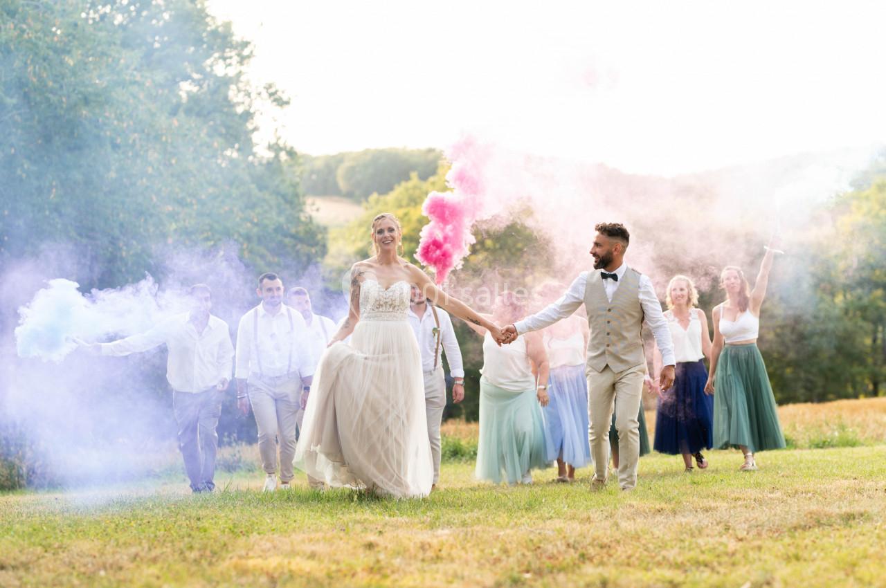 Un couple de mariés marche dans un champ, suivi d'invités tenant des fumigènes colorés.