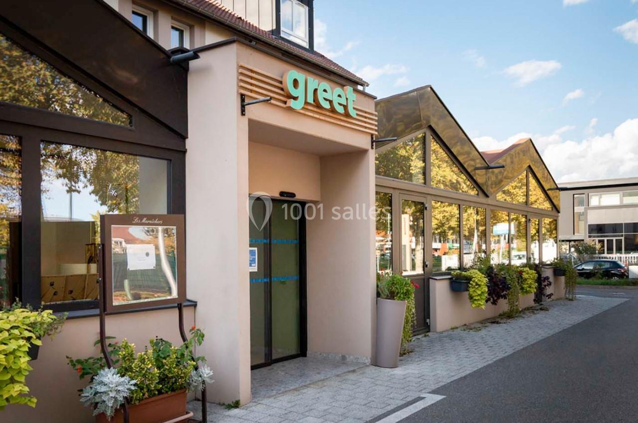 Entrée d'un bâtiment avec une enseigne ’greet’, entourée de plantes et de grandes baies vitrées.