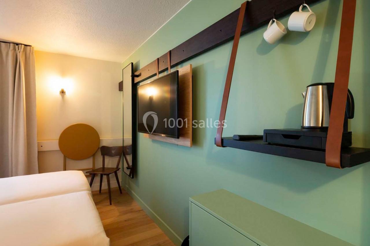 Chambre moderne avec deux lits, mur vert, télévision murale, bouilloire sur étagère et lumière douce.