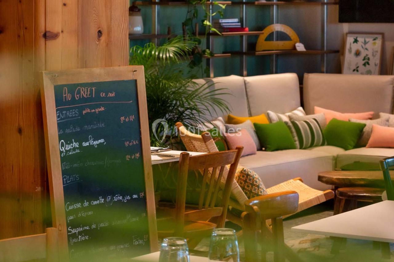 Intérieur d'un café chaleureux avec une ardoise de menu, des fauteuils en bois et un canapé entouré de plantes.