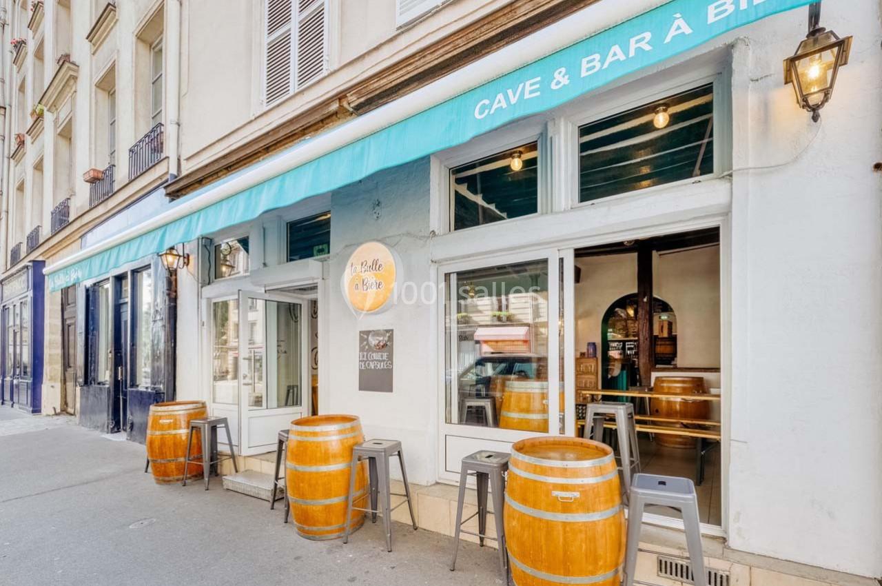 Façade d'un bar avec terrasse, tabourets et tonneaux en bois, situé le long d'une rue urbaine.