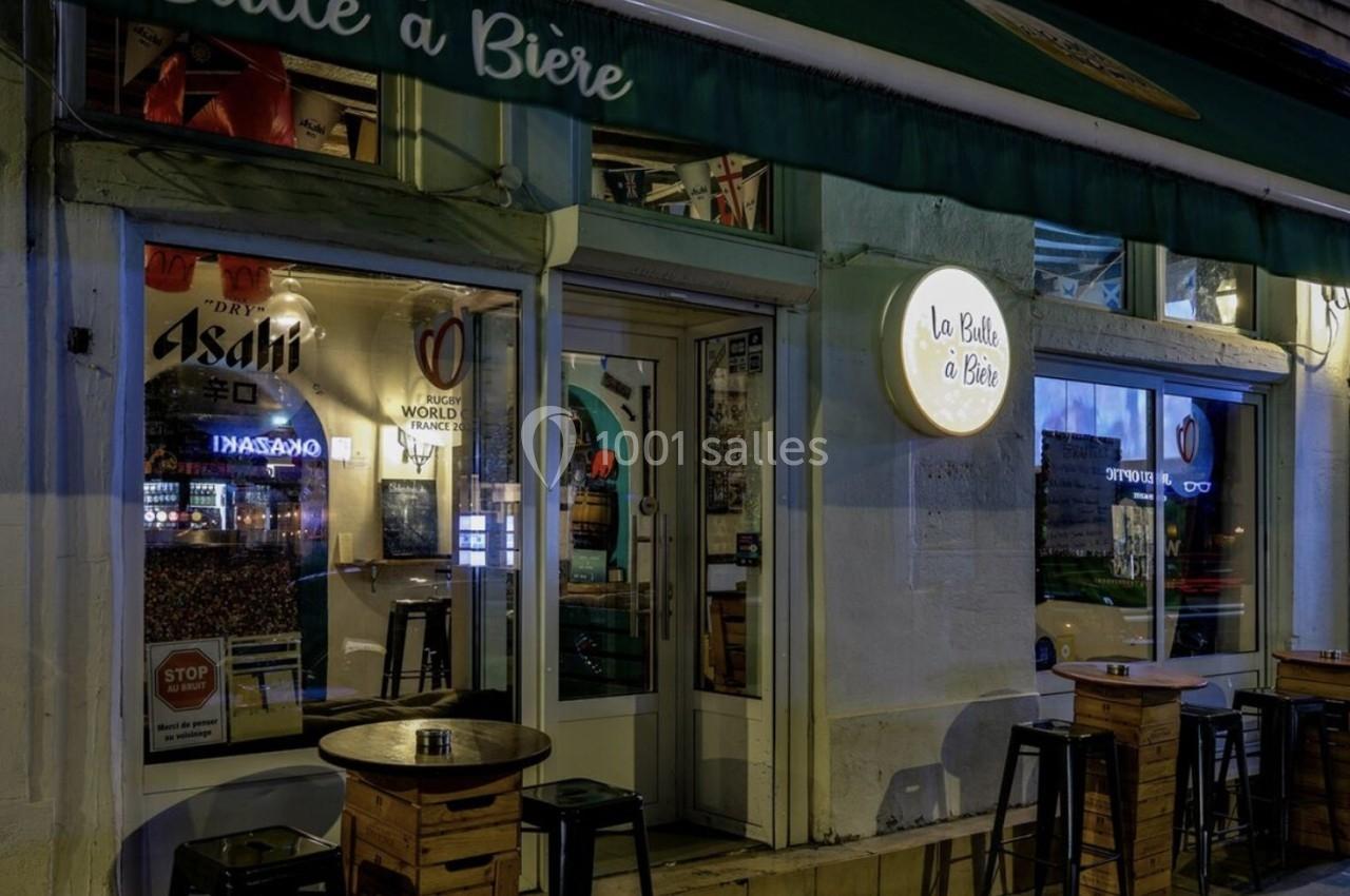 Façade d'un bar éclairé de nuit avec des tables hautes en bois disposées sur le trottoir.