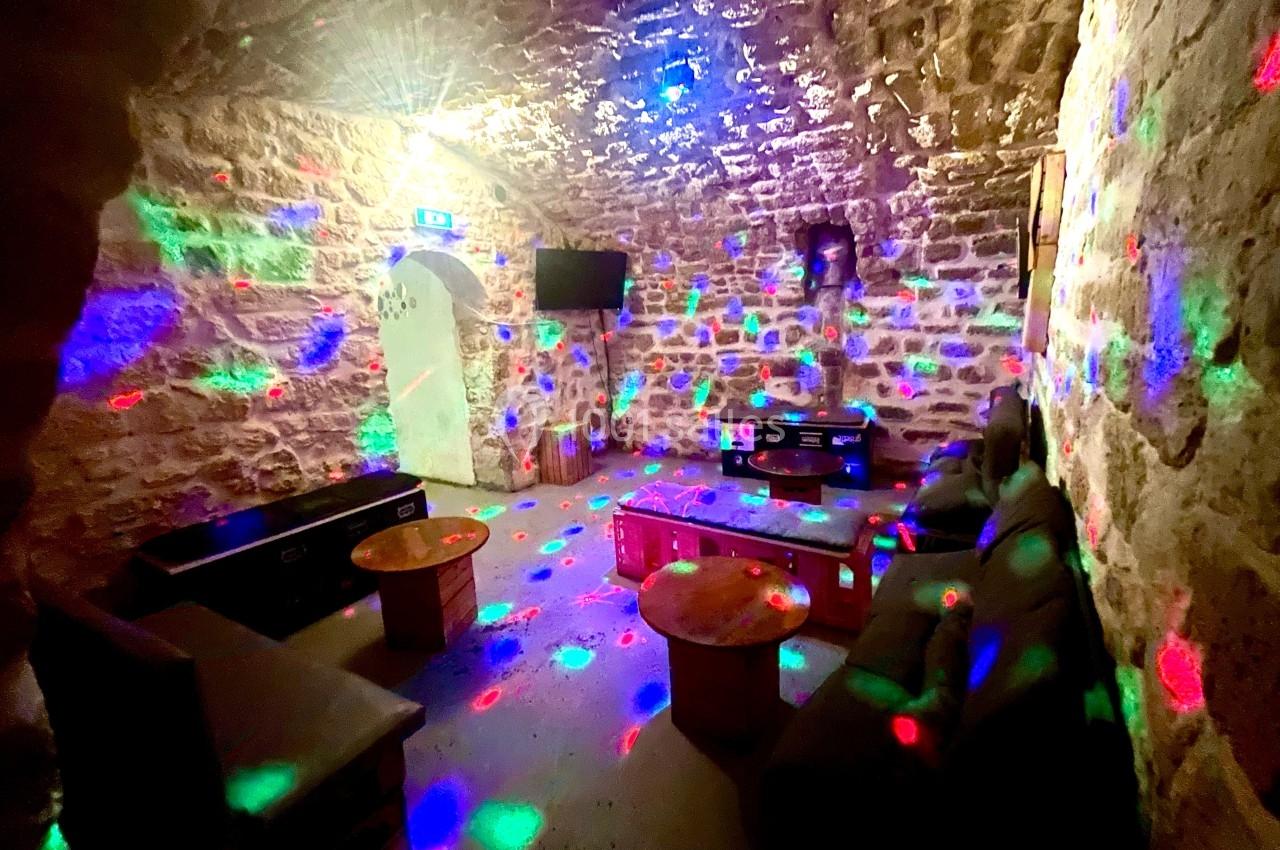 Salle voûtée en pierre avec éclairage coloré, canapés, tables basses et écran mural.